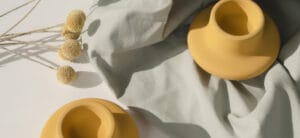 Two yellow ceramic vases on a light gray fabric, with three spherical dried flowers on the side, casting soft shadows on a white surface.