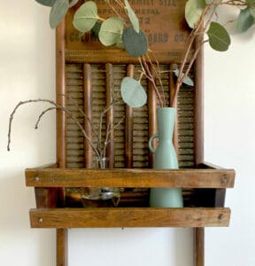 A wooden washboard mounted on a wall, with a small shelf holding a glass jar and a blue vase containing branches and leaves.