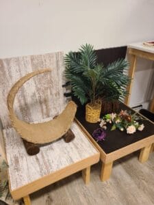 A rustic chair shaped like a crescent moon is next to a wooden table adorned with a potted plant and a variety of decorative flowers.