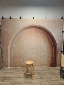 A wooden stool sits on a wood floor in front of a backdrop with an arched design.