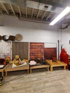 A room with wooden platforms displaying various props, including a teddy bear, chairs, rugs, and a basket against a backdrop of wood and brick patterns.