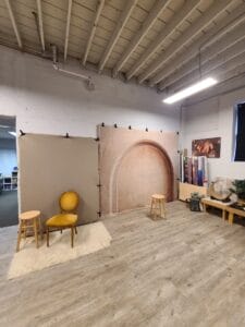 Photo studio setup with a vintage yellow chair, two stools, and backdrop featuring an arched design. Wooden floor, rug, and assorted props in a well-lit room.