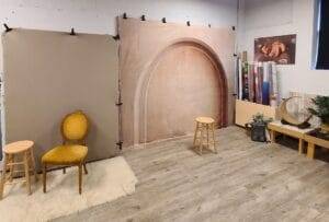 Photography studio setup with a beige backdrop, two stools, a vintage chair on a rug, and photography equipment including rolls of background paper and lighting accessories.