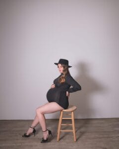 A pregnant woman wearing a black dress, high heels, and a hat sits sideways on a wooden stool against a plain background.