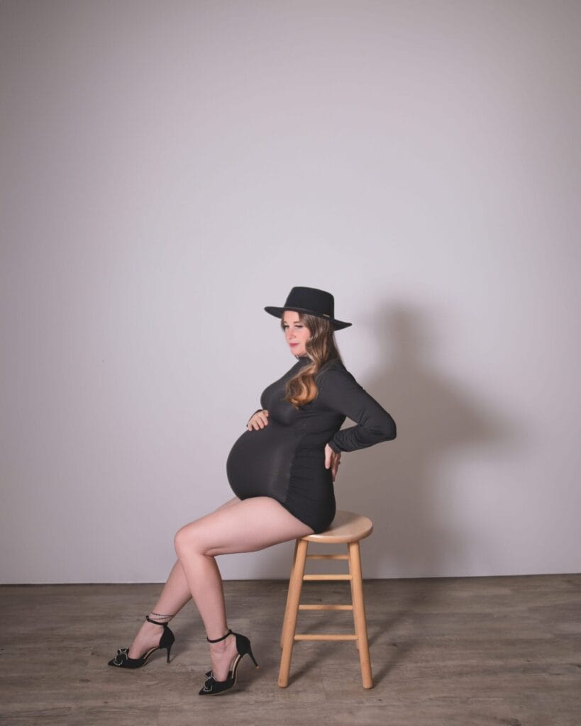 A pregnant woman wearing a black dress, high heels, and a hat sits sideways on a wooden stool against a plain background.