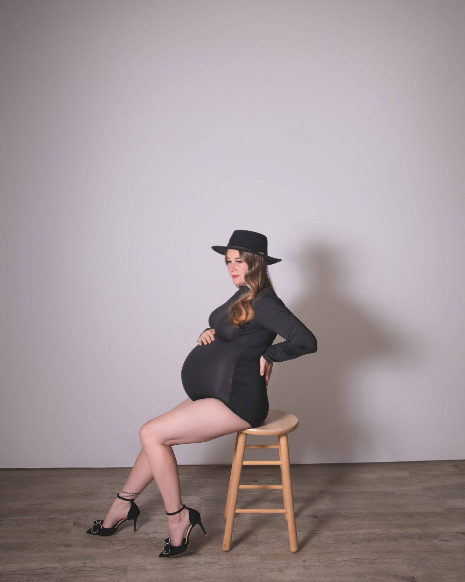 A pregnant woman wearing a black dress, high heels, and a hat sits sideways on a wooden stool against a plain background.
