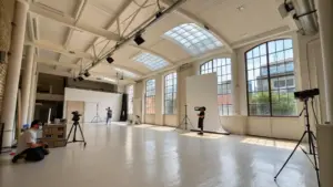 Spacious photo studio with high ceilings, large windows, and professional lighting equipment. Two photographers and a model are positioned for a shoot.