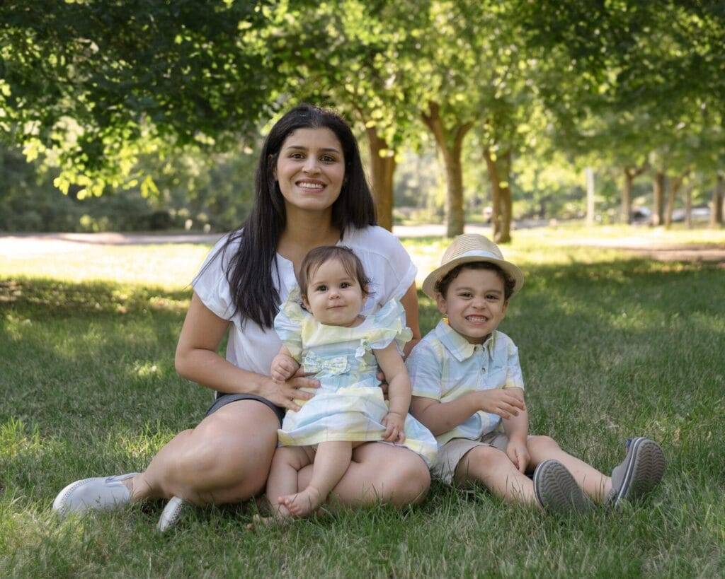 A woman sits on grass with two young children, a toddler and a slightly older boy in a hat, all smiling in a park setting with trees in the background.