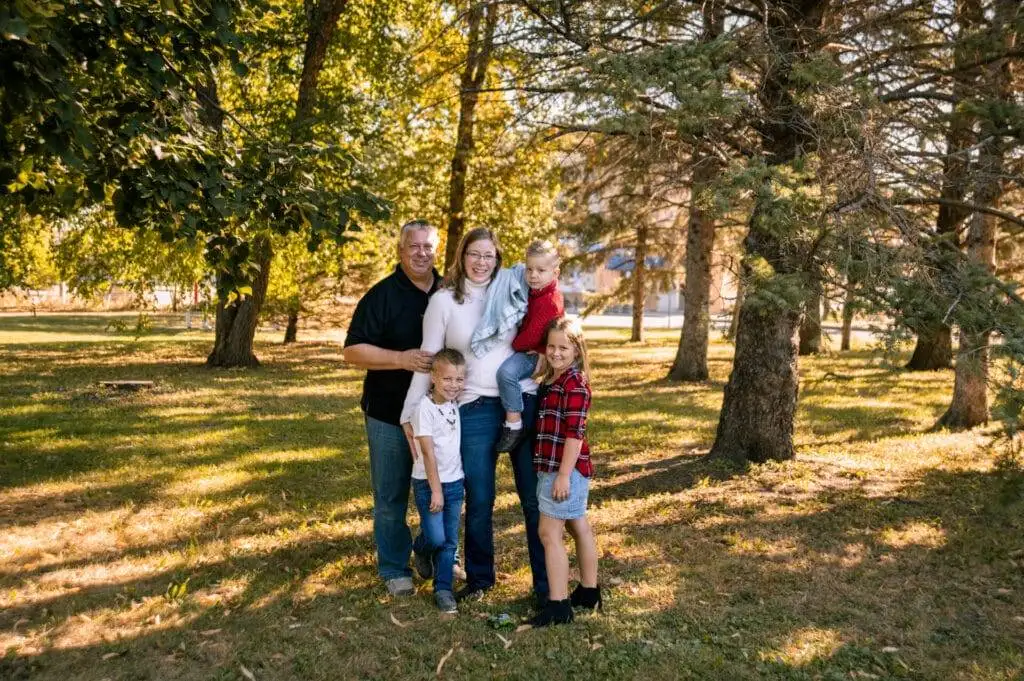 A family of five poses together outdoors in a wooded area, standing on grass with trees and sunlight in the background.