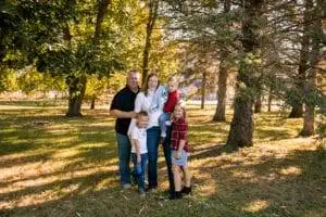 A family of five poses together outdoors in a wooded area, standing on grass with trees and sunlight in the background.