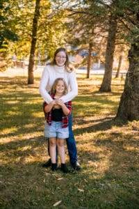 An adult and a child stand together smiling in a sunlit park, with trees and grass in the background. The adult is standing behind the child, arms around her shoulders.