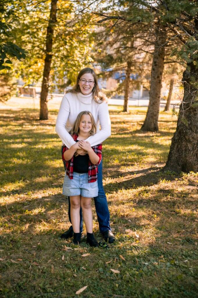 An adult and a child stand together smiling in a sunlit park, with trees and grass in the background. The adult is standing behind the child, arms around her shoulders.