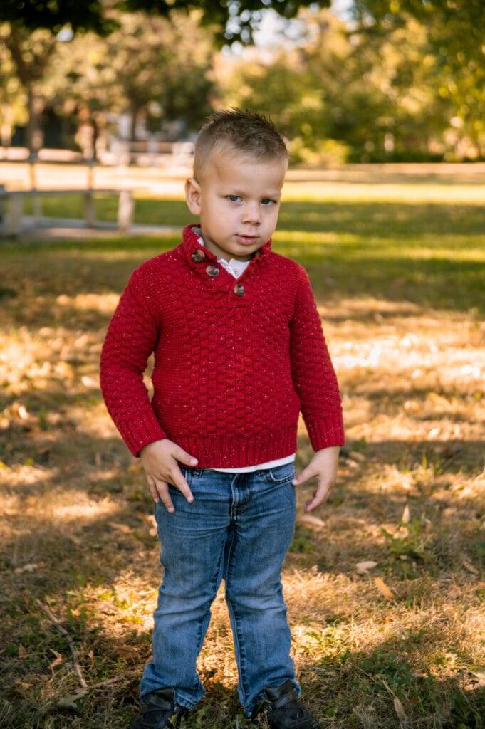 A young child in a red sweater and blue jeans stands on grass in a park with trees and sunlight in the background.