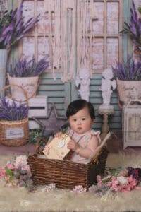 A baby sits in a wicker basket holding a wooden sign, surrounded by flowers, with a floral and rustic backdrop featuring baskets and decorative items.