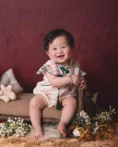 A smiling baby in a white floral outfit sits on a cushion surrounded by flowers and foliage with a burgundy backdrop.