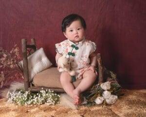 A toddler in a floral outfit sits on a small wooden bench, holding a stuffed toy, surrounded by flowers and cushions against a maroon backdrop.