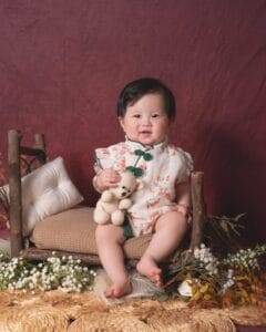 A baby sits on a rustic wooden bench, holding a small stuffed bear, surrounded by flowers and cushions, against a maroon backdrop.