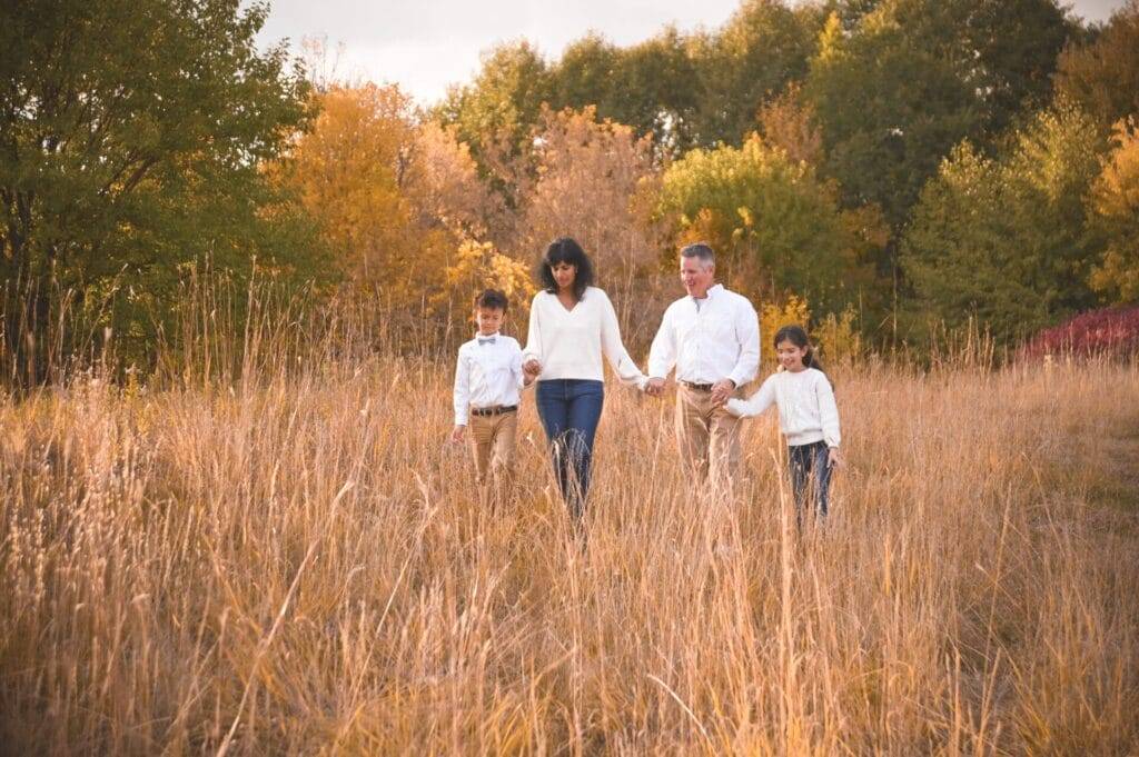 A family of four walks through a grassy field with autumn trees in the background, all wearing light-colored tops and tan pants.