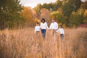 A family of four walks through a grassy field with autumn trees in the background, all wearing light-colored tops and tan pants.