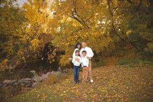 A family of four stands together on a leaf-covered ground near a small river, surrounded by autumn trees with yellow leaves.