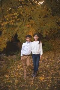 Two children stand side by side outdoors on fallen leaves, with autumn trees in the background. The boy wears a white shirt and khaki pants; the girl wears a white sweater and jeans.
