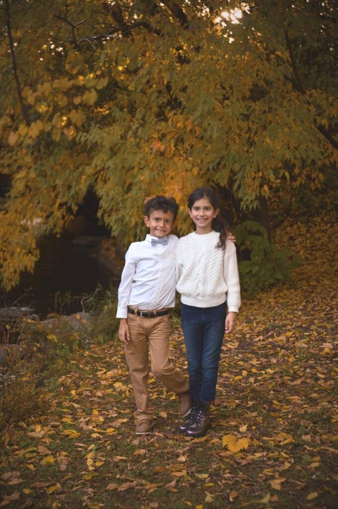 Two children stand side by side outdoors on fallen leaves, with autumn trees in the background. The boy wears a white shirt and khaki pants; the girl wears a white sweater and jeans.