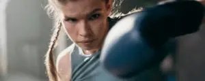 A woman with braided hair wearing boxing gloves throws a punch, focused on her training in a gym setting.