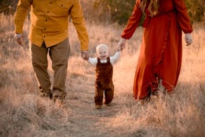 A small child walks through a dry grassy field holding hands with two adults, all dressed in autumn-colored clothing.
