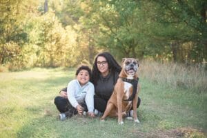 A woman, a young boy, and a brown and white dog sit together on grass in a sunlit park with trees in the background.