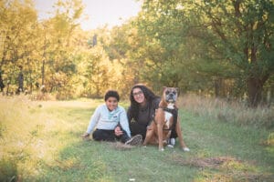 A woman, a child, and a brown dog sit on grass in a sunlit park with trees in the background.