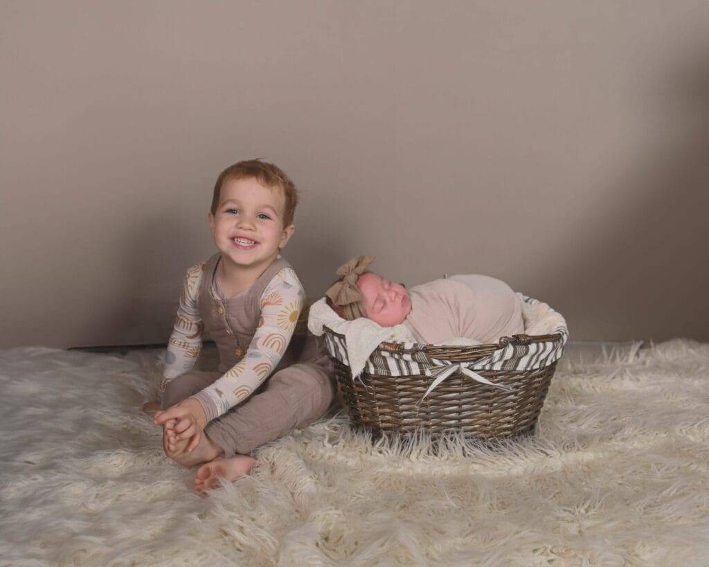 A young child sits smiling on a white rug next to a newborn baby wrapped in a blanket, lying in a wicker basket.