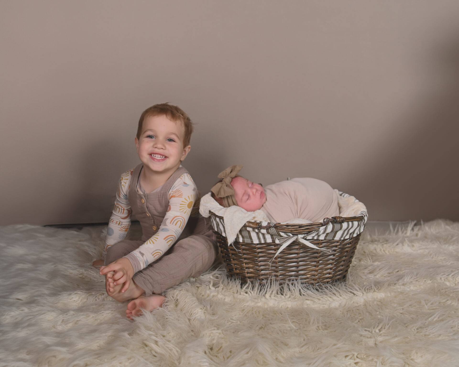 A young child sits smiling on a white rug next to a newborn baby wrapped in a blanket, lying in a wicker basket.
