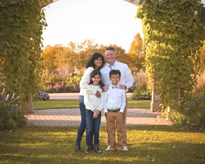 A family of four poses together outdoors under a leafy archway, with autumn trees and garden plants in the background.