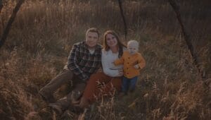 A man, woman, and toddler pose together outdoors in a grassy field, sitting and standing among tall dry grass under soft evening light.
