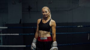 A female boxer wearing gloves, a black sports top, and red-black shorts stands in a boxing ring, looking at the camera.