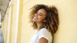 A woman with curly hair wearing a white t-shirt leans against a yellow wall, smiling and looking slightly upward.