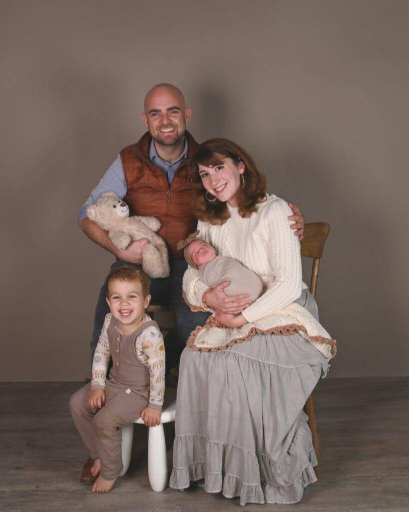 A family portrait with two adults, a young child holding a teddy bear, and a baby wrapped in a blanket sitting on the mother’s lap, all posing indoors against a plain backdrop.