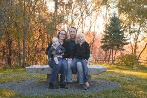 A family of four sits on a stone bench outdoors in a park with autumn trees in the background, posing for a photo.