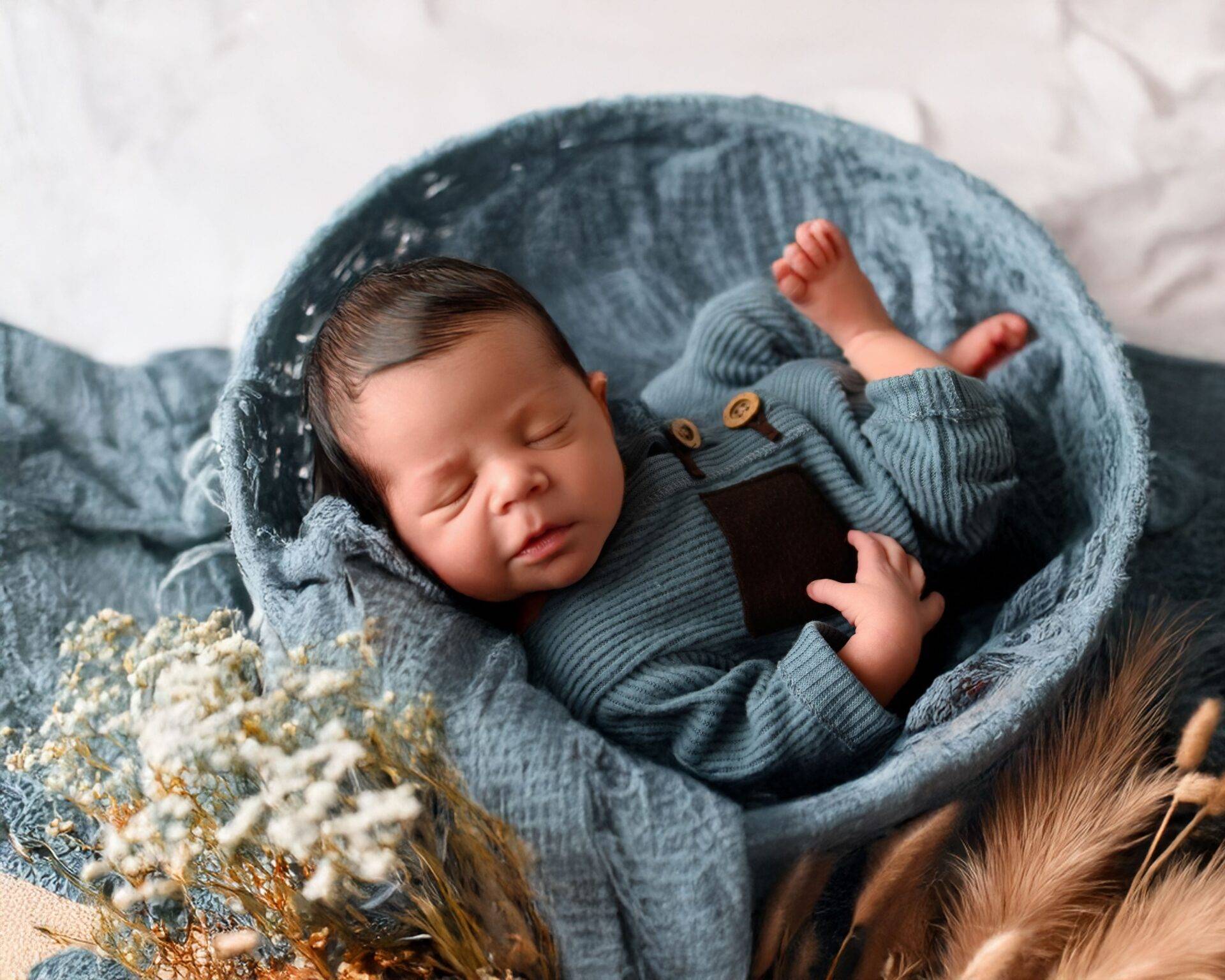 Newborn baby sleeping on a blue blanket inside a round basket, wearing a blue knitted outfit; dried flowers are placed nearby.