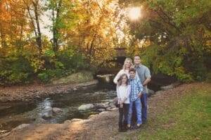 A family of four stands near a creek with autumn trees and a stone bridge in the background; sunlight filters through the foliage.