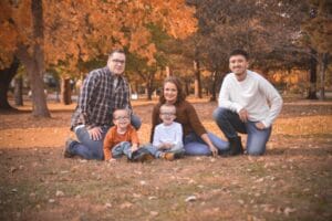 A family of five poses together on the ground in a park with autumn foliage in the background.