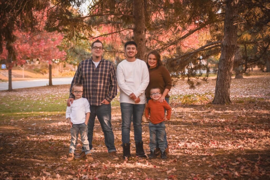 A family of five poses outdoors among autumn trees and fallen leaves, with three children standing in front of two adults.