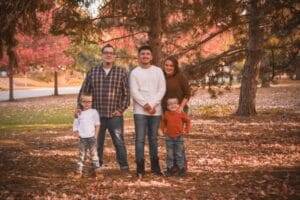 A family of five poses outdoors among autumn trees and fallen leaves, with three children standing in front of two adults.
