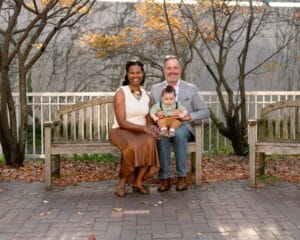 A woman, man, and baby sit together on a wooden bench outdoors in autumn, with fallen leaves and bare trees in the background.
