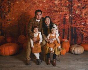A family of four poses together indoors with autumn-themed decorations, including pumpkins and fall foliage in the background.