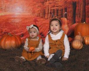 Two young children sit on dark soil surrounded by pumpkins, wearing fall-themed outfits, with an autumn forest backdrop.