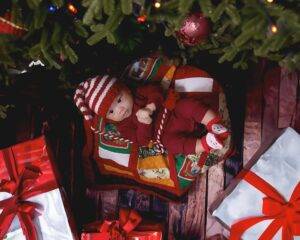 A baby in a red outfit and striped hat lies on a quilt under a Christmas tree, surrounded by wrapped gifts with red ribbons.