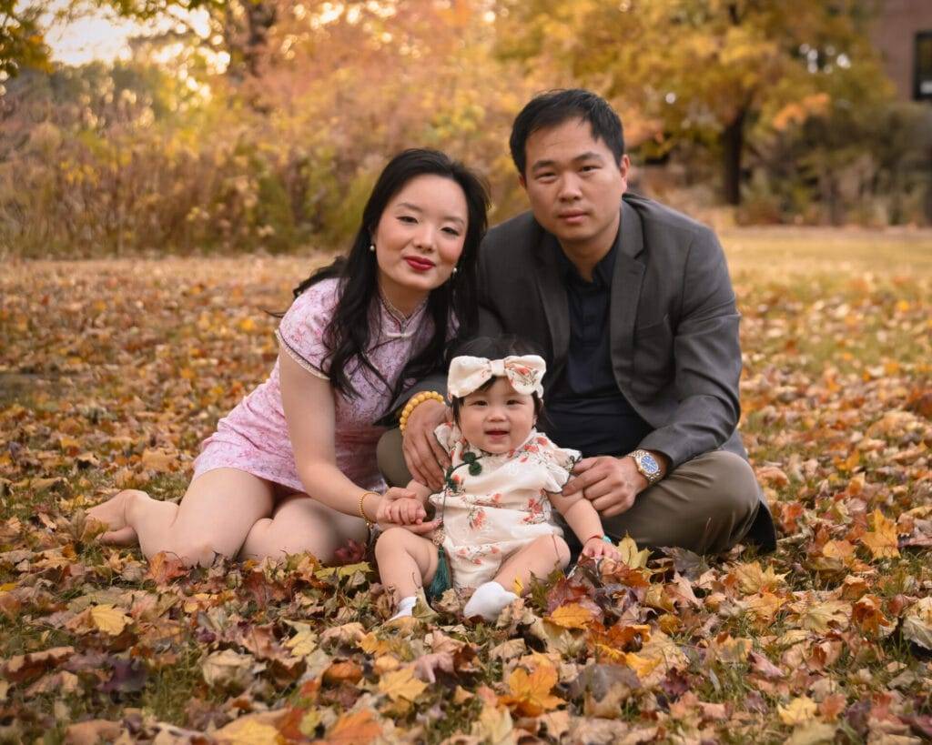 An adult woman, an adult man, and a baby sit together on autumn leaves in a park, with trees and foliage in the background.