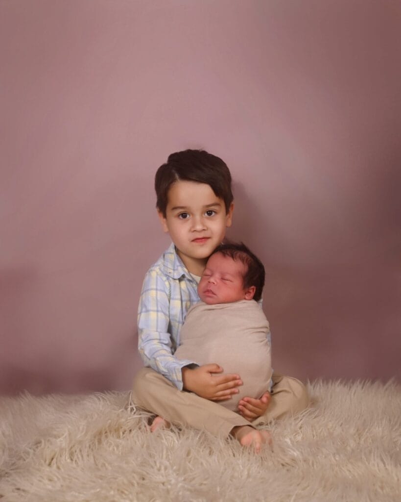 A young boy sits on a furry rug, holding a swaddled newborn baby in his lap, both posed against a plain mauve background.