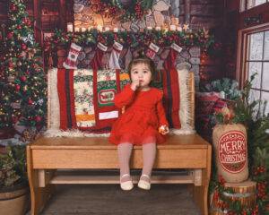 A young child in a red dress sits on a wooden bench in a festive, Christmas-themed room with stockings, a tree, and holiday decorations.